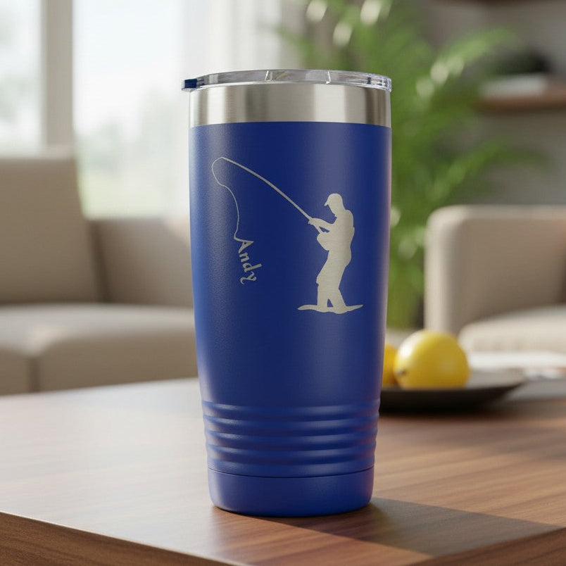 A royal blue insulated tumbler with a silver stainless steel lid sits on a wooden table in a modern living room setting. The tumbler features an etched laser-engraved design of a fisherman and the name 'Andy'.