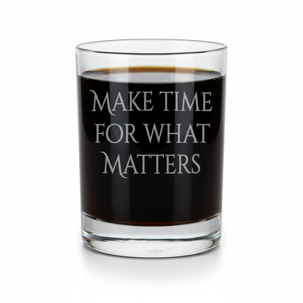 A clear rocks glass with the phrase 'MAKE TIME FOR WHAT MATTERS' etched on the front, filled with a dark amber whiskey, set on a light wood surface against a white background.