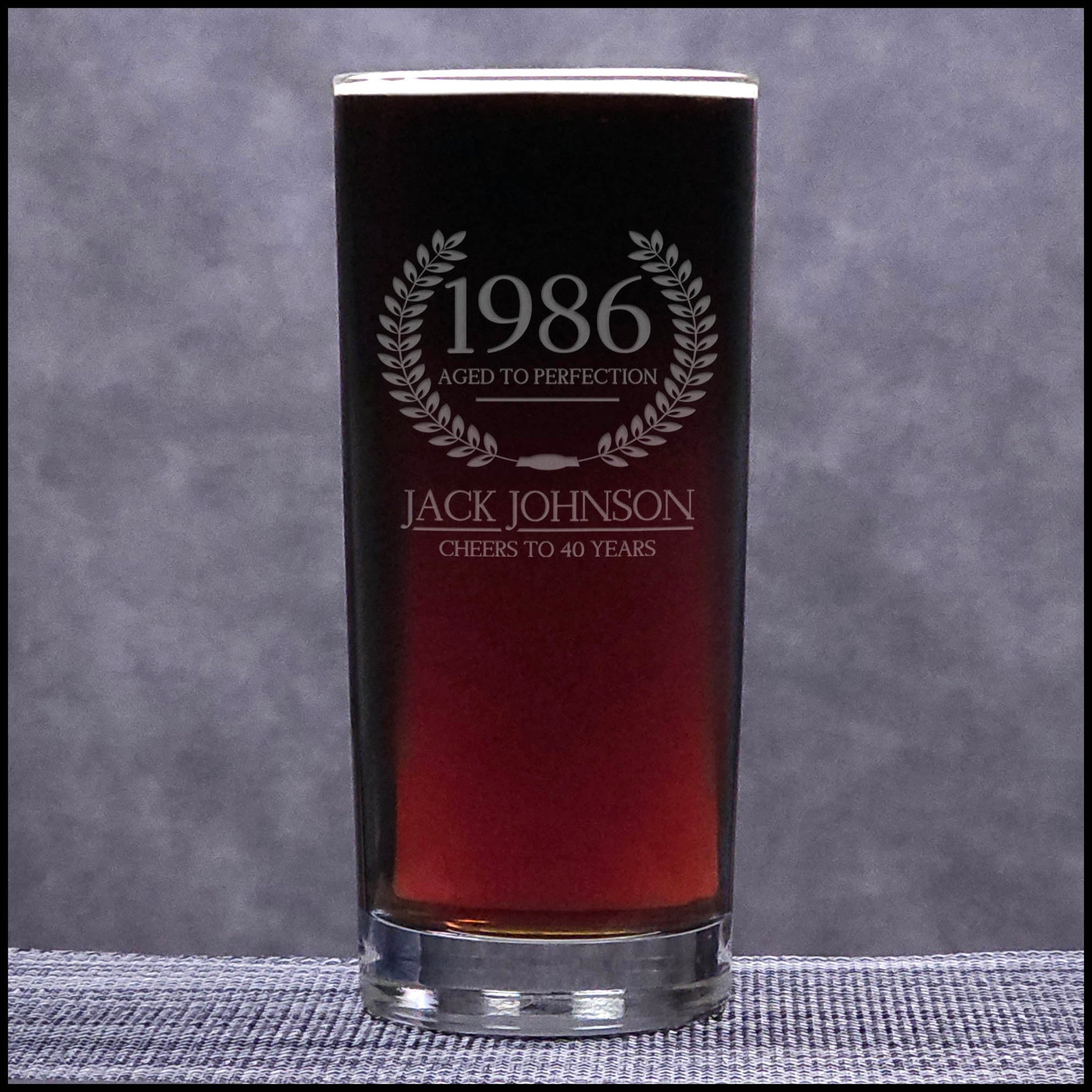 Glass with engraved text '1986 Aged to Perfection Jack Johnson Cheers to 40 Years' on a gray background