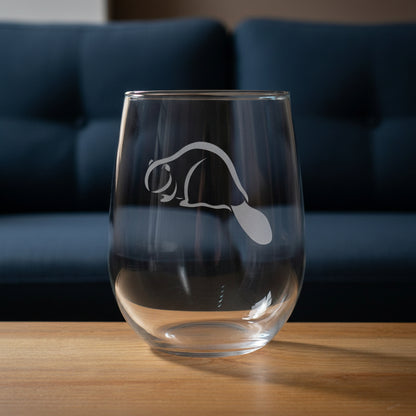 A clear stemless wine glass with a gray beaver silhouette graphic on the front, resting on a wooden surface against a blue couch.