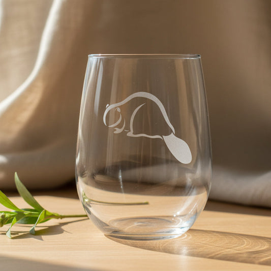 A clear stemless wine glass with an etched beaver design on the front, resting on a wooden surface 