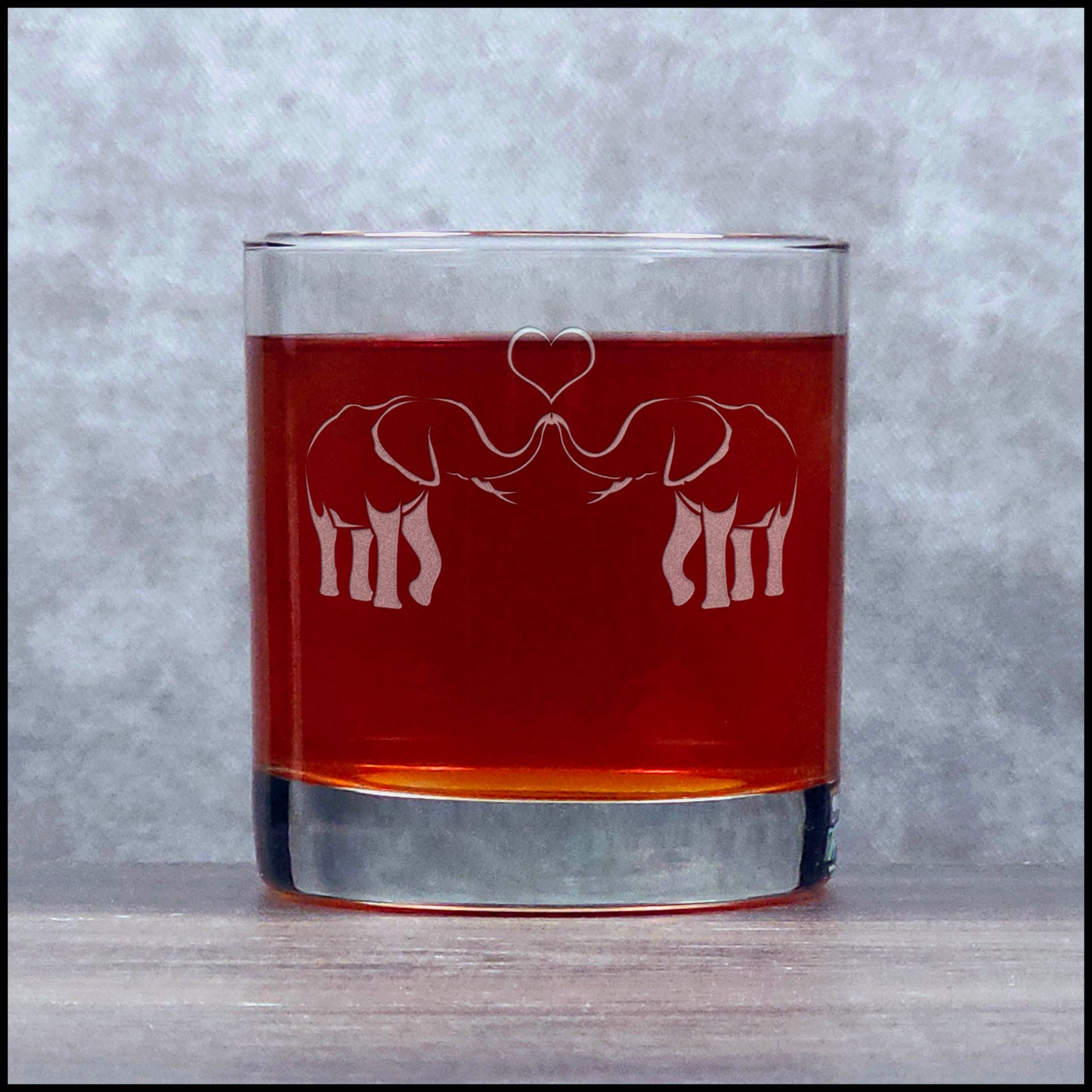 A clear glass rocks tumbler with an etched design of two elephants facing each other with a heart-shaped trunk touch, shown filled with an amber-red liquid against a green couch