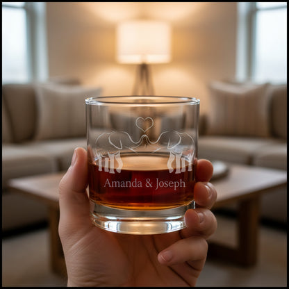 A clear rocks glass filled with amber-red liquid, featuring an etched design of two elephants facing each other with a heart above and the names 'Amanda & Joseph' below, set against a tliving room.