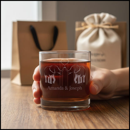 A hand holds a clear rocks glass filled with amber-brown liquid, etched with an elephant design and the names 'Amanda & Joseph'. In the softly blurred background, a kraft paper gift bag and a cream drawstring pouch sit on a wooden table.