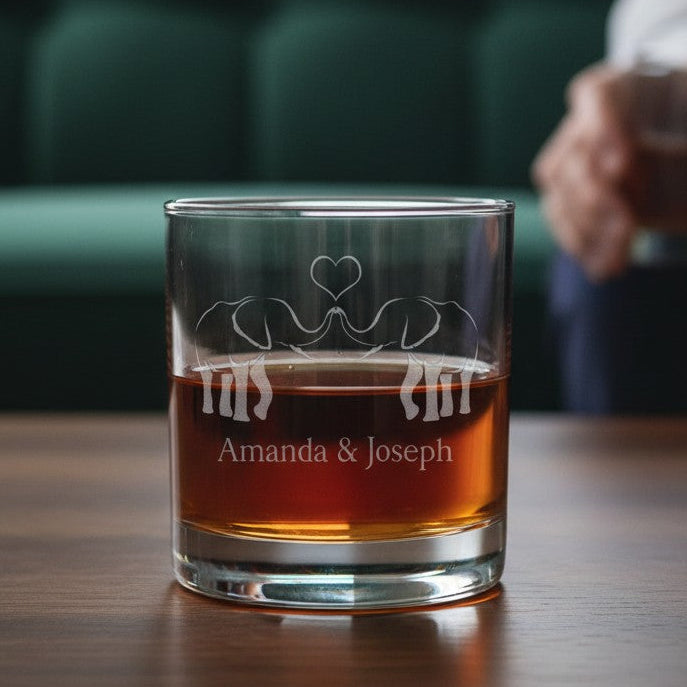 A clear whiskey tumbler with an etched elephant and heart design, filled with amber liquor, sits on a wooden table. The background shows a blurred green sofa and a person holding another glass.