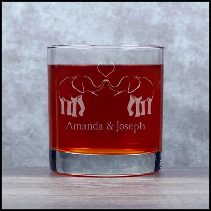 A clear rocks glass filled with amber-red liquid, featuring an etched design of two elephants facing each other with a heart above and the names 'Amanda & Joseph' below, set against a white background.