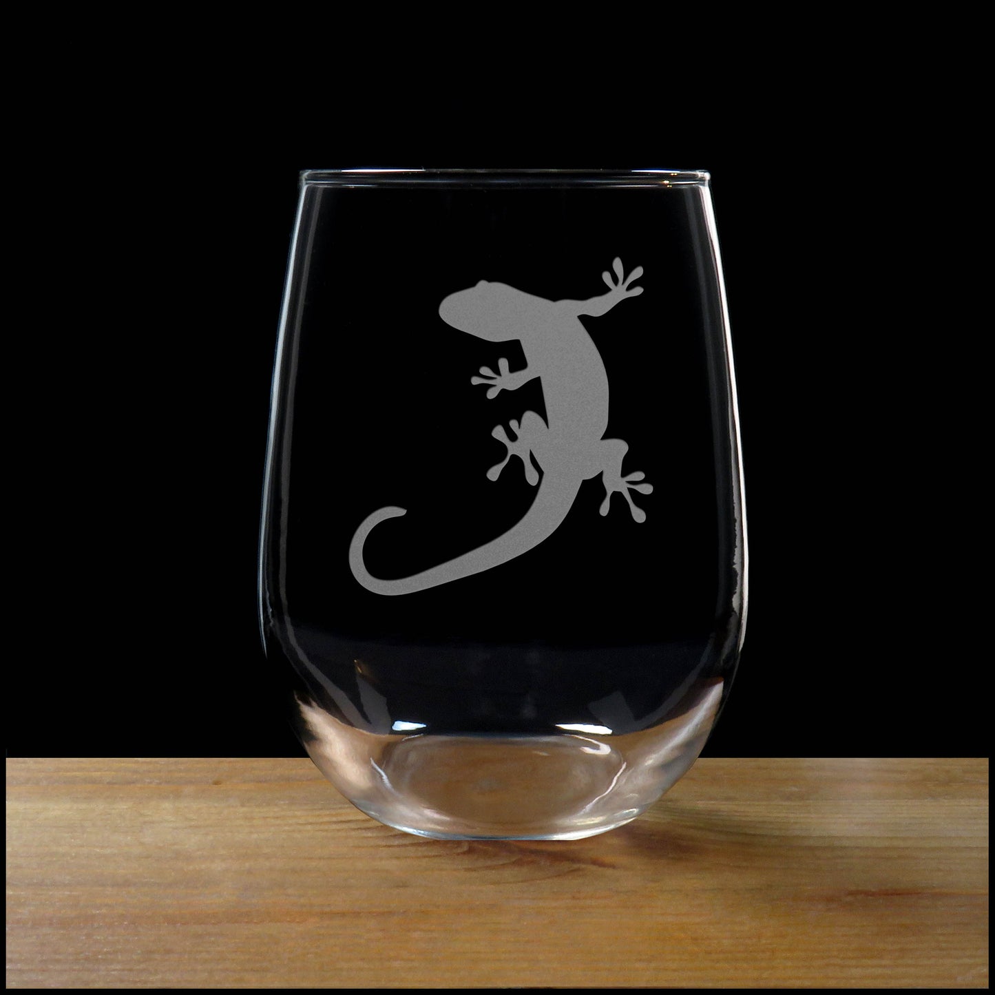 A clear stemless wine glass with an etched image of a gecko on the glass resting on a wooden surface against a solid black background.