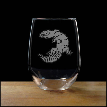 A clear stemless wine glass with an etched image of a gecko on the glass resting on a wooden surface against a solid black background.