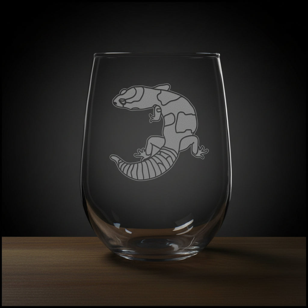 A clear stemless wine glass with an etched image of a gecko resting on a wooden surface against a solid black background.