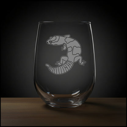 A clear stemless wine glass with an etched image of a gecko resting on a wooden surface against a solid black background.
