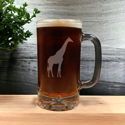 A clear glass beer mug with a handle, filled with an amber drink and topped with white foam, featuring a white giraffe silhouette design on the front. The mug sits on a light wood surface against a gray wood-plank background with a green plant visible on the left.