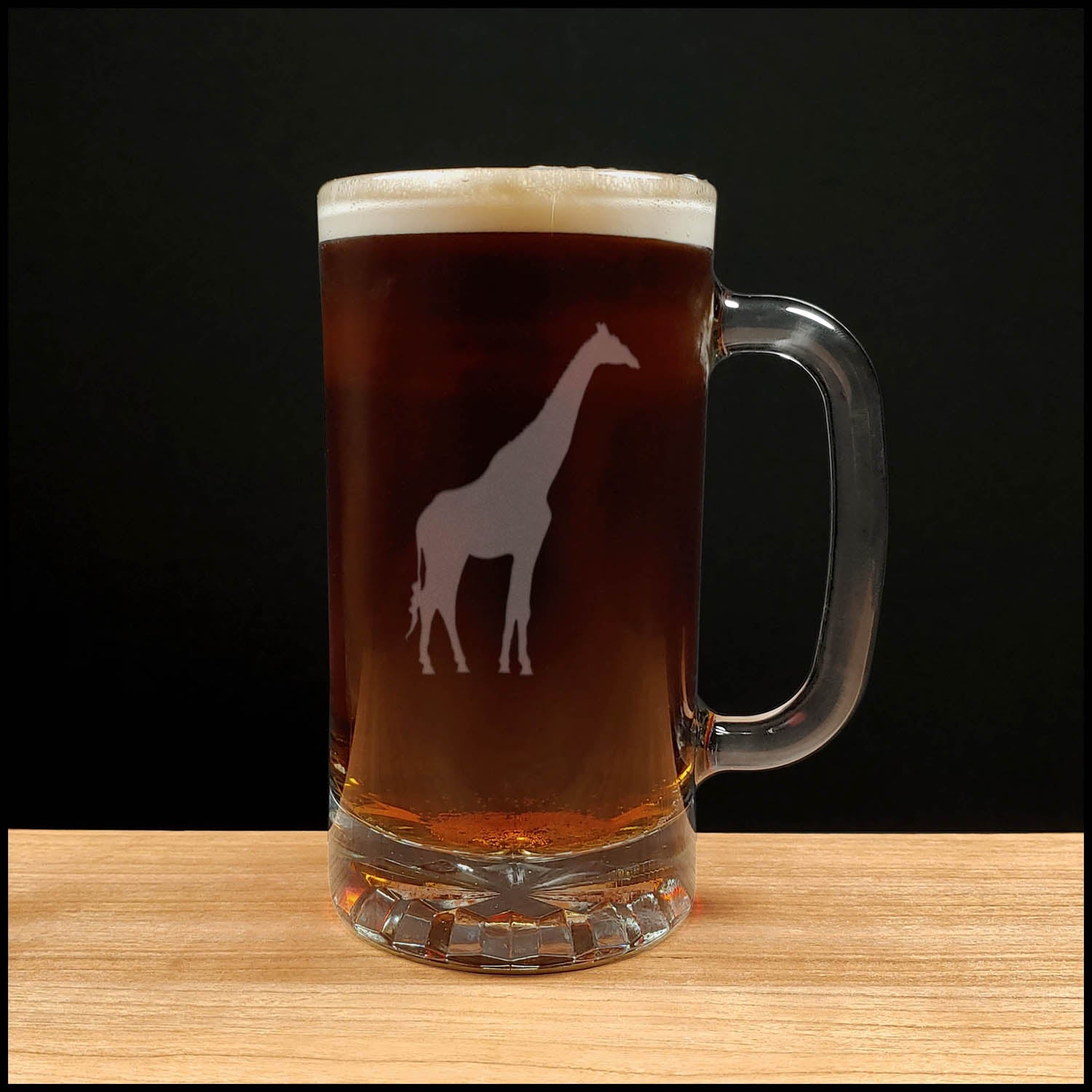 A clear glass beer mug with a handle, filled with dark beer and topped with white foam, featuring a white giraffe silhouette design on the front, set on a light wood surface against a black background.