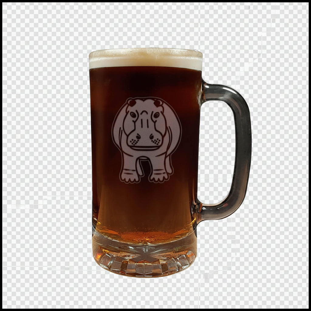A clear glass beer mug with a handle, filled with dark amber beer and topped with white foam, featuring a etched hippo design on the front, set on a light wood surface against a white background.