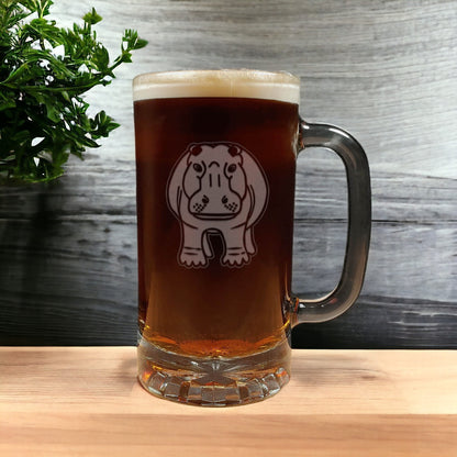 A clear glass beer mug filled with dark amber beer and topped with white foam, featuring a cartoon hippo graphic on the front, set on a wooden surface against a gray wood plank background with a green plant at the side.