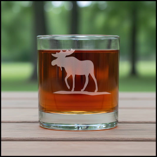 A clear rocks glass tumbler with an etched moose silhouette, filled with amber-brown liquid, set against trees  in the background.