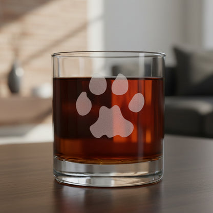 A clear rocks glass filled with an amber-red liquid, featuring a frosted paw print design on the front, set on a dark wooden surface with a living room background.