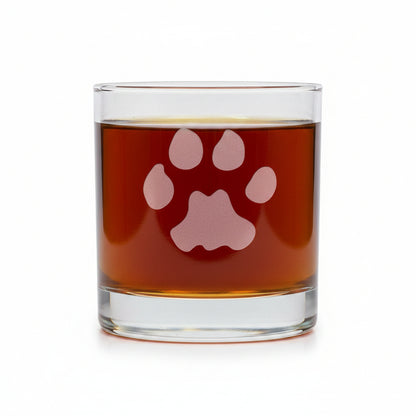 A clear glass rocks tumbler with an etched paw print design, filled with a dark reddish-brown liquid, set on a reflective gray surface against a white background.