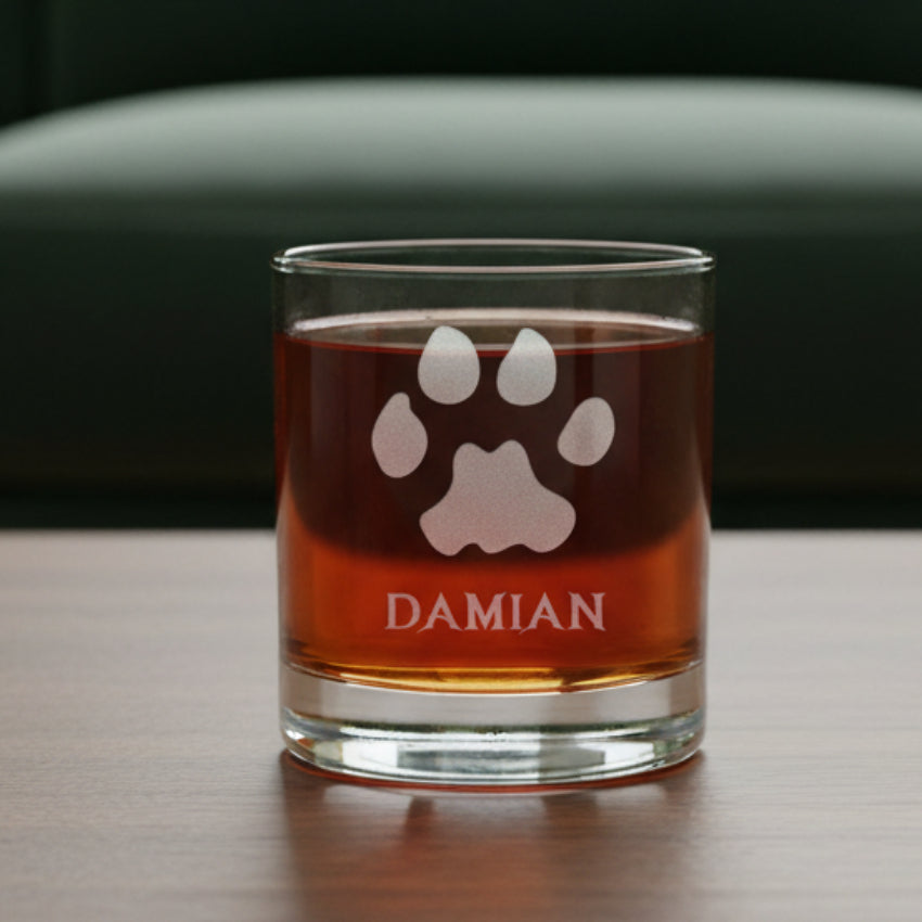 A clear glass rocks tumbler with a custom etched paw print and the name 'DAMIAN', filled with an amber-colored liquid, sitting on a dark wooden surface with a green couch background.