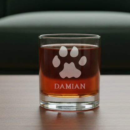 A clear glass rocks tumbler with a custom etched paw print and the name 'DAMIAN', filled with an amber-colored liquid, sitting on a dark wooden surface with a green couch background.