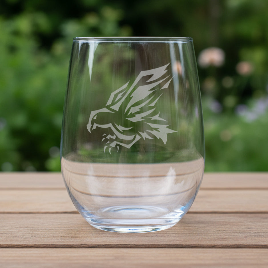 A clear stemless wine glass with a white eagle head graphic printed on the front, resting on a wooden surface against a garden background.