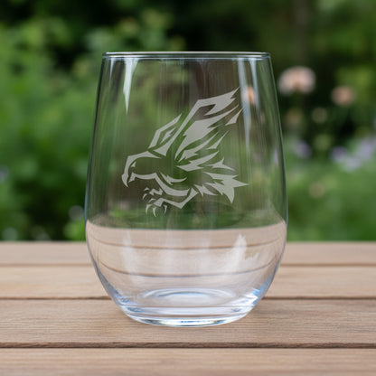 A clear stemless wine glass with a white eagle head graphic printed on the front, resting on a wooden surface against a garden background.