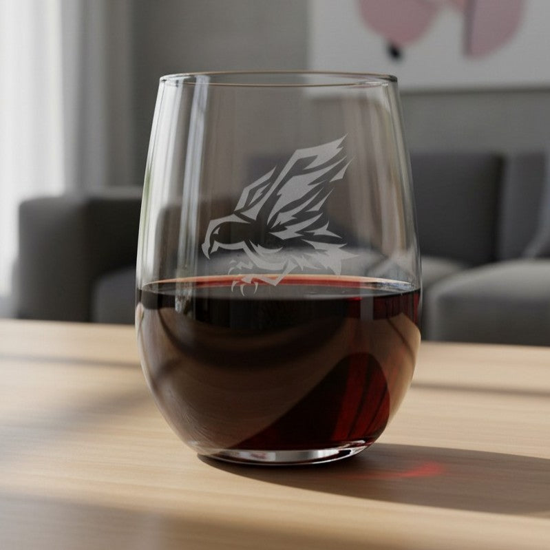 A clear stemless wine glass with an etched eagle design, partially filled with red wine, sitting on a light wooden table in a blurred living room setting.