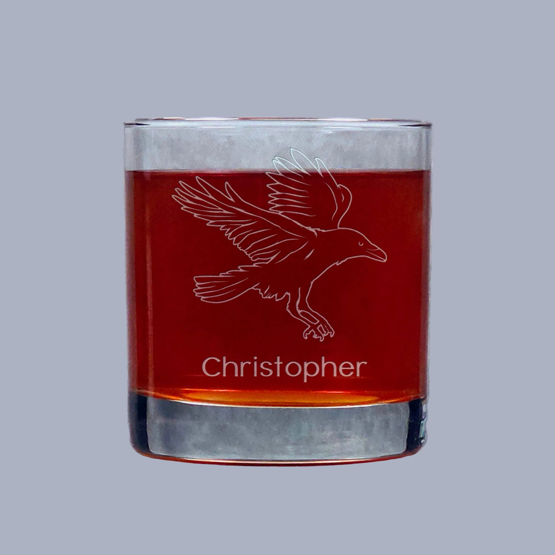 Glass tumbler with engraved Raven design and 'Christopher' text, filled with a amber red liquid on a gray background.