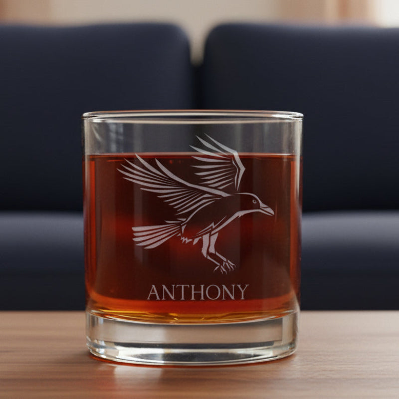 Glass tumbler with engraved Raven design and 'Anthony' text, filled with a red liquid on a wooden surface.