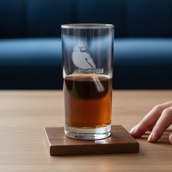 A clear glass tumbler with a white bird illustration sits on a square wooden coaster on a light wood table, with a hand resting nearby and a blue tufted sofa in the background.