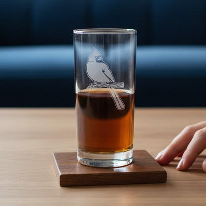 A clear glass tumbler with a white bird illustration sits on a square wooden coaster on a light wood table, with a hand resting nearby and a blue tufted sofa in the background.