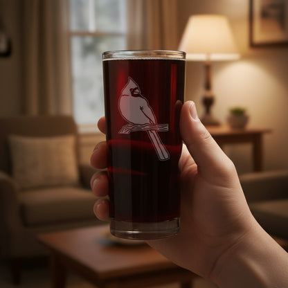 A hand holds a clear glass tumbler with a white cardinal perched on a branch, filled with a very dark red beverage, set against a softly blurred living room background with a lamp and sofa.