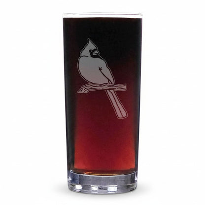 A clear highball glass with a gradient of dark brown to reddish-brown liquid inside, featuring a white etched cardinal perched on a branch on the front, set against a plain white background.