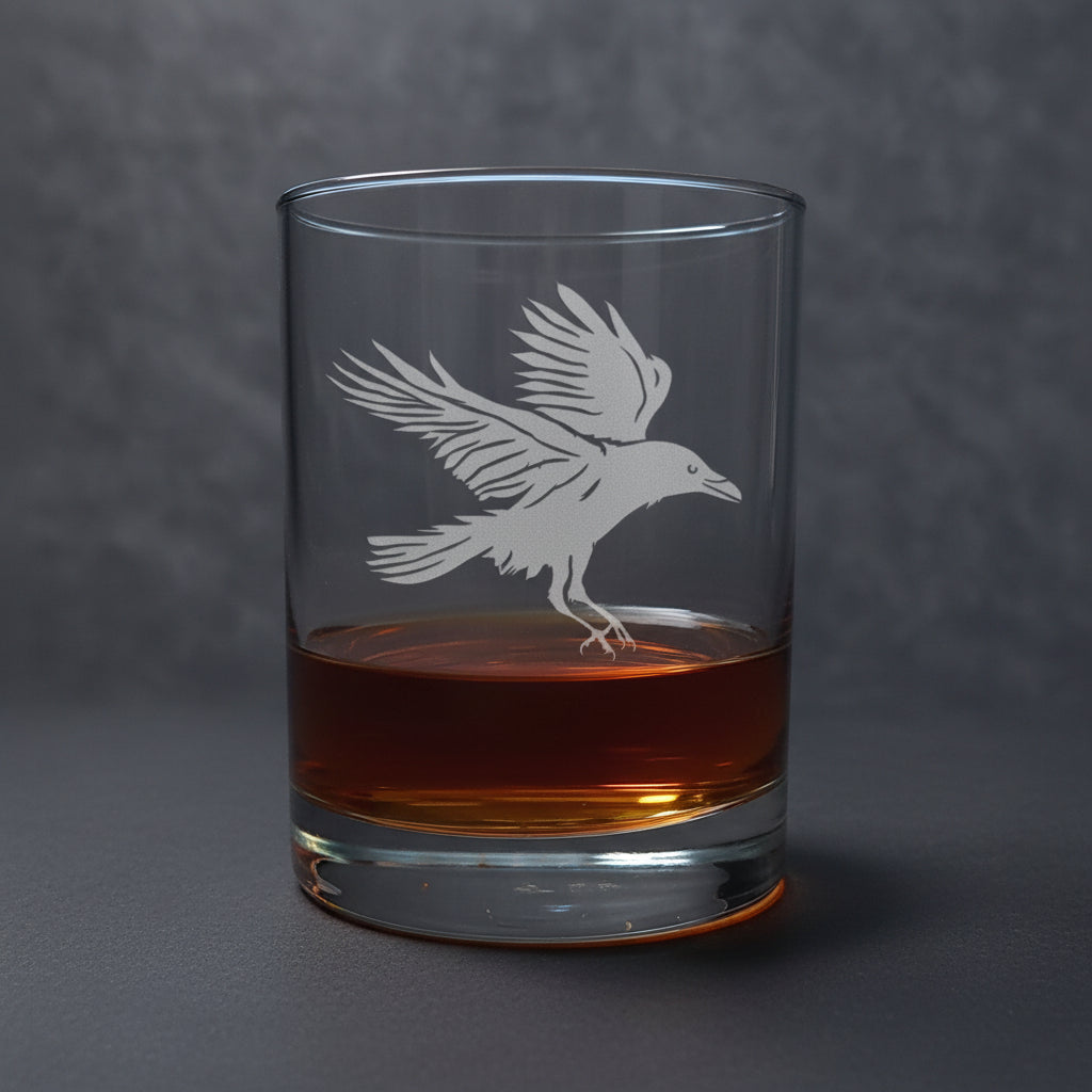 Clear glass tumbler with raven etching filled with amber liquid on a dark background