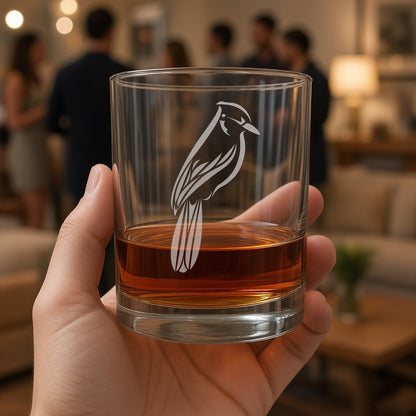 A hand holds a clear glass with an etched  bluejay, with amber whiskey, against a softly blurred indoor social setting with people in the background.