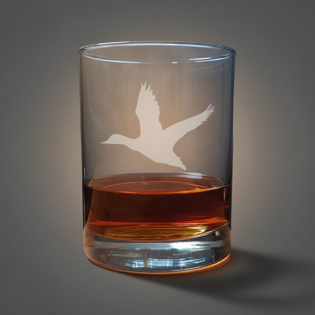Clear glass tumbler with a bird etching filled with amber liquid with a grey background
