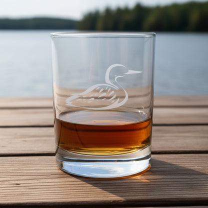 A clear glass rocks tumbler with an etched loon design, partially filled with amber whiskey, set on a wooden surface against lake and trees in the background.