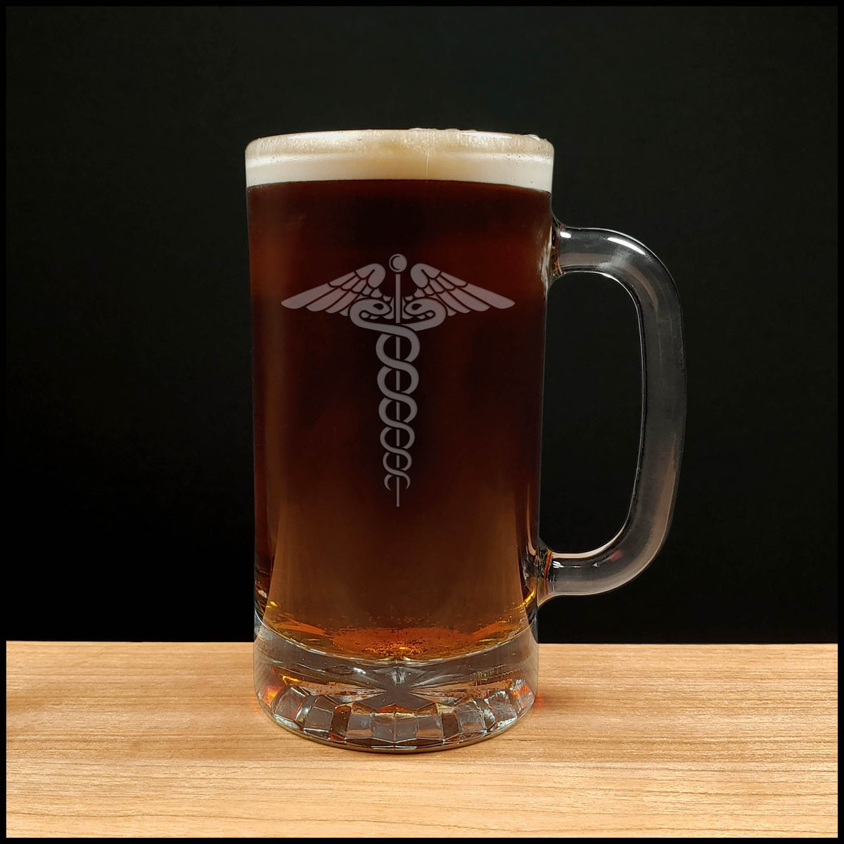 A clear glass beer mug filled with dark amber beer and topped with white foam, featuring an etched medical caduceus symbol on the front, set on a light wood surface against a black background.