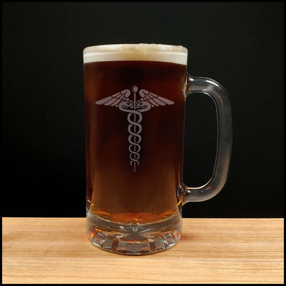 A clear glass beer mug filled with dark amber beer and topped with white foam, featuring an etched medical caduceus symbol on the front, set on a light wood surface against a black background.