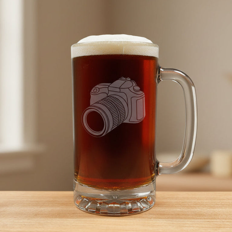 A clear glass beer mug filled with dark beer, featuring an etched camera design on the front, set on a light wood 