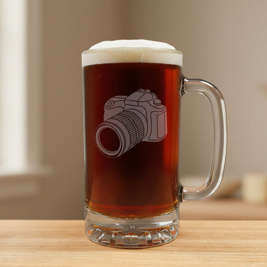 A clear glass beer mug filled with dark beer, featuring an etched camera design on the front, set on a light wood 