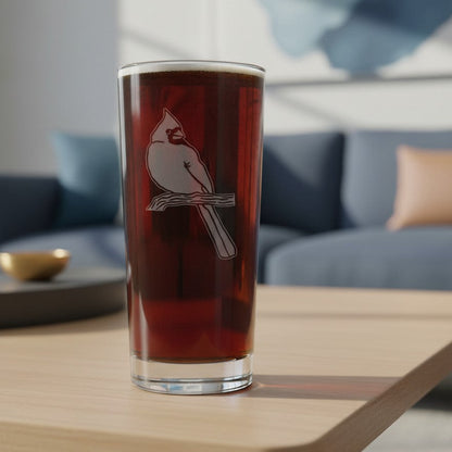 A clear pint glass with a white cardinal perched on a branch, filled with a dark reddish-brown beverage and set on a light wood table in a modern living room with a blue sofa in the background.