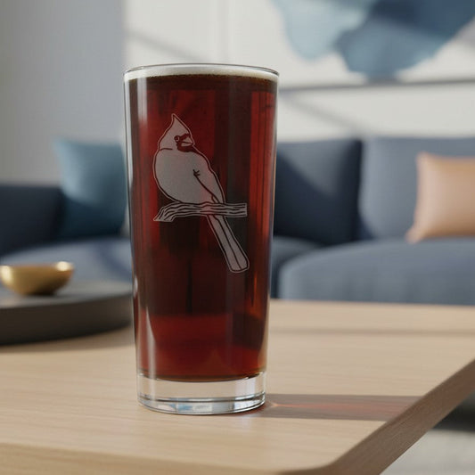 A clear pint glass with a white cardinal perched on a branch, filled with a dark reddish-brown beverage and set on a light wood table in a modern living room with a blue sofa in the background.