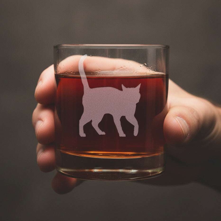 A clear rocks glass filled with amber-red liquid, with a hand holding it. Featuring a etched cat silhouette design on the front, set against a textured gray background.