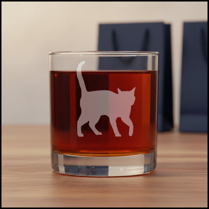 A clear rocks glass filled with amber-red liquid, featuring a etched cat silhouette design on the front, set against two gift bags