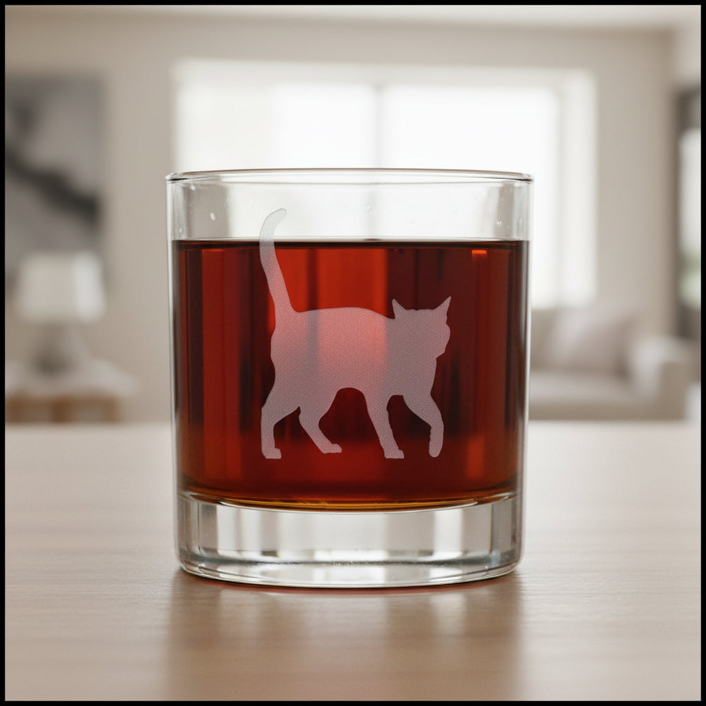 A clear rocks glass filled with amber-red liquid, featuring a fetched cat silhouette design on the front, set against a modern living room background.