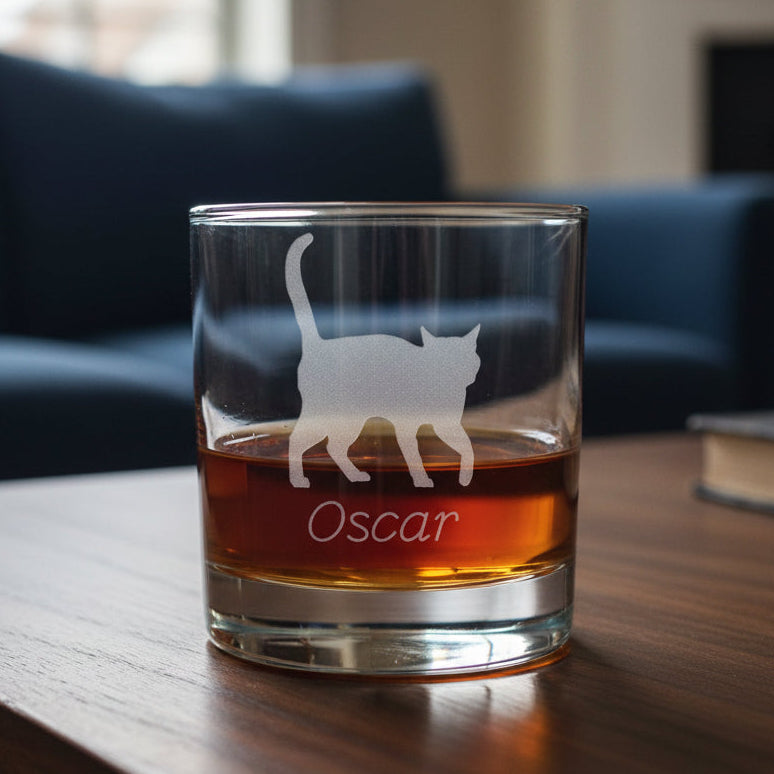A clear glass rocks tumbler filled with an amber-red drink, featuring an etched silhouette of a walking cat and the name 'Oscar' on the front, set against a blue couch background.