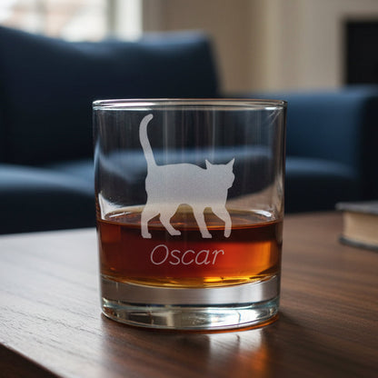 A clear glass rocks tumbler filled with an amber-red drink, featuring an etched silhouette of a walking cat and the name 'Oscar' on the front, set against a blue couch background.