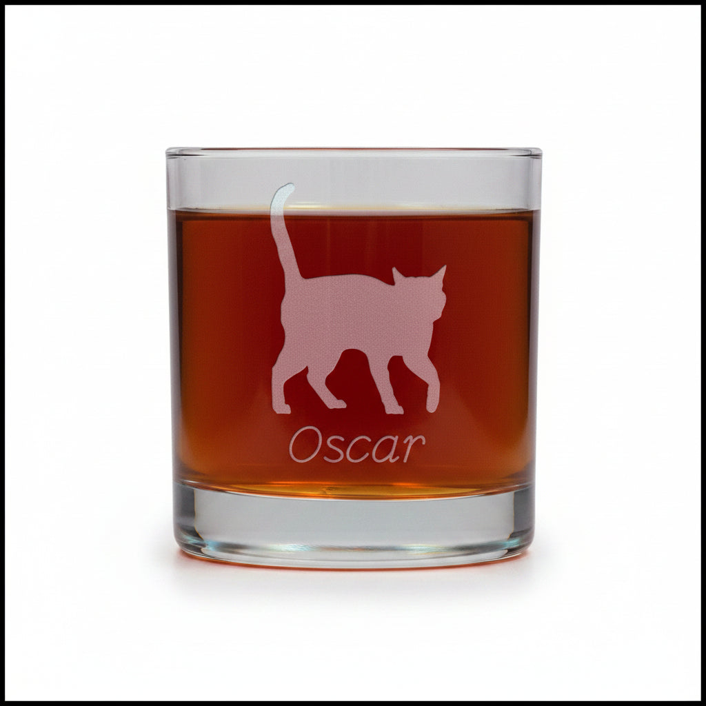 A clear glass rocks tumbler filled with an amber-red drink, featuring an etched silhouette of a walking cat and the name 'Oscar' on the front, set against a white  background.