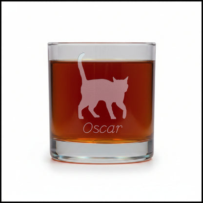 A clear glass rocks tumbler filled with an amber-red drink, featuring an etched silhouette of a walking cat and the name 'Oscar' on the front, set against a white  background.