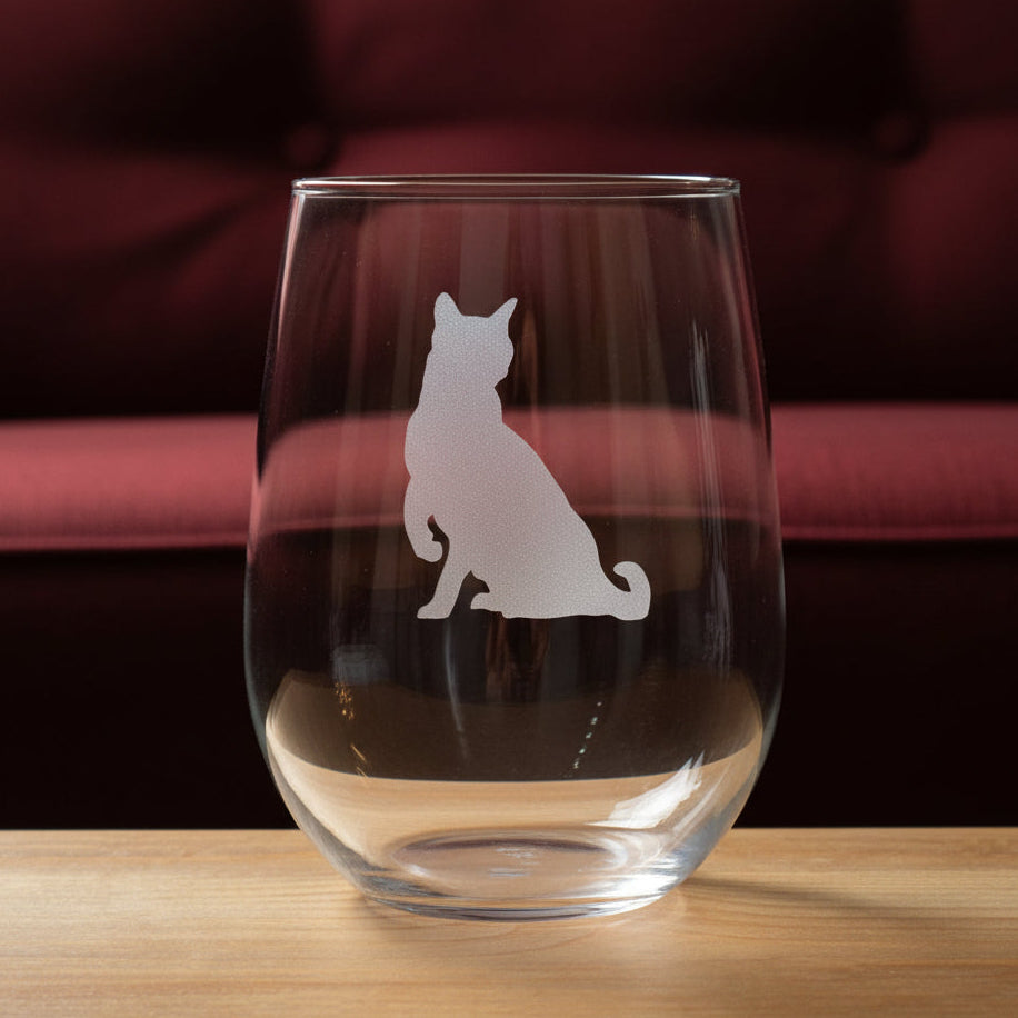 A clear stemless wine glass with a gray etched silhouette of a sitting cat, displayed on a wooden surface against a red couch.
